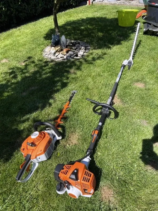 Zwei orange STIHL Heckenscheren nebeneinander auf einer Rasenfläche abgelegt: eine kompakte Motor-Heckenschere und eine Langarm-Heckenschere mit ausgefahrenem Schaft