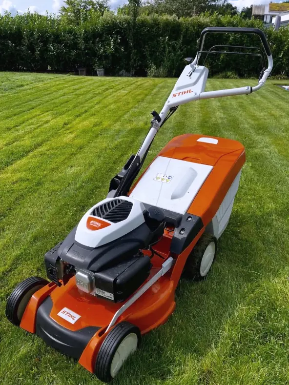 Orange-weißer STIHL Benzin-Rasenmäher mit angebautem Grasfangkorb auf frisch gemähtem Rasen, im Hintergrund die gemähten Bahnen und eine Hecke